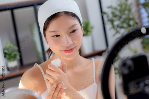Young asian woman presenting and review cosmetic product with on social media, beauty influencer reviewing skincare cream product on hand with phone, live streaming or broadcast, content creator.