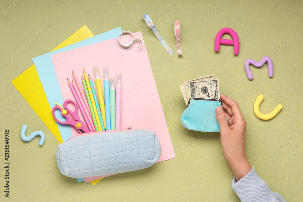 Fototapeta premium Childs hand holding a coin purse with money next to school supplies and colorful paper on a green background, a childs hand holds a light blue coin purse filled with hundred dollar bills, financial