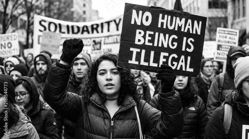 A young woman with a raised fist holds a sign stating 'NO HUMAN BEING IS ILLGAL' during a black and white protest march with many people in the background.