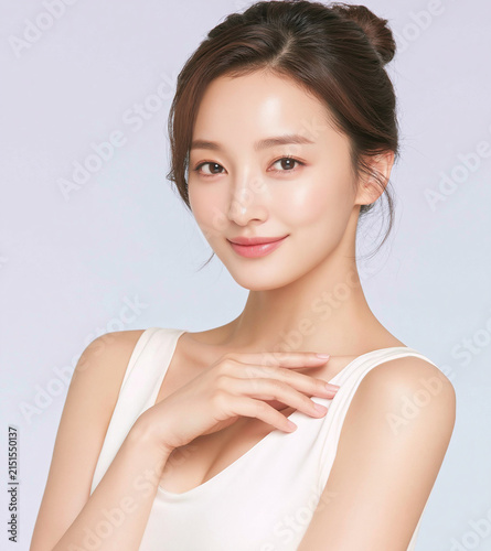 Vertical headshot portrait of elegant young East Asian woman touching collarbone with smooth glowing skin, soft makeup, and updo hair on light background