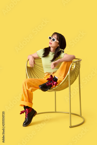Stylish young Asian woman with bows in armchair on yellow background