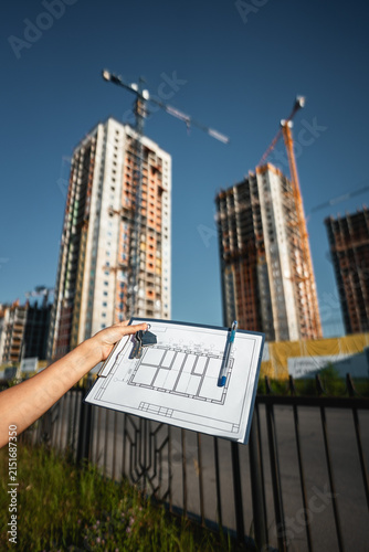 Engineer reviews architectural drawings while standing beside active construction site with cranes and residential buildings. Professional planning, project management and quality inspection concept.