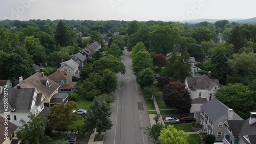 Tree-lined suburban street in New Jersey neighborhood reveals colonial-style homes with steep gabled roofs, wraparound porches, brick chimneys and manicured green lawns. Aerial drone descend shot