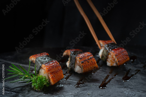 Chopsticks are picking up a piece of Japanese sushi with grilled eel on top, beautifully arranged on a black stone tray. Traditional Japanese culinary concept.