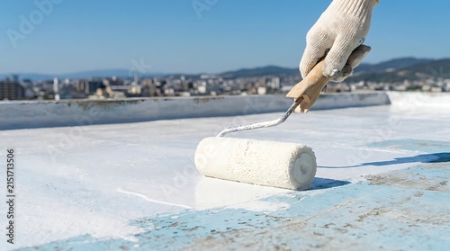 A close-up view of a hand in a white glove using a paint roller to apply a thick white waterproof sealant onto a flat roof under a blue sky