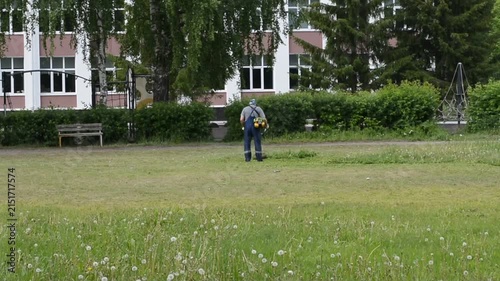 Gardener mowing green lawn with trimmer near building. Worker trimming grass in park area. Outdoor landscaping maintenance service by professional groundskeeper.