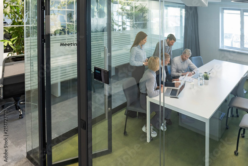 A view through a glass partition shows a modern, busy office interior. Business people are working at desk