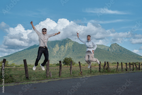 A joyful Asian couple laugh and jump on a rural road with a green Baluran mountain landscape and blue sky behind them, capturing adventure, freedom, travel and outdoor happiness during a scenic day tr