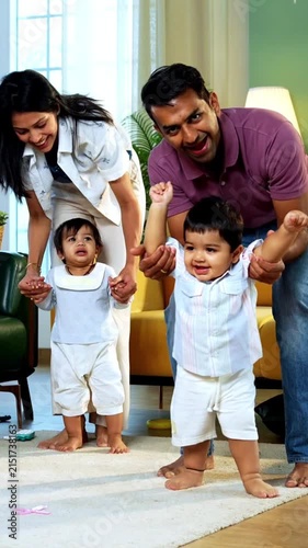 Indian Asian young parents helping twin infant babies walk by holding their hands inside modern home living room, guiding first steps together as smiling babies enjoy playful family bonding moment