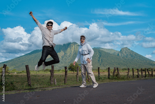 A joyful Asian couple laugh and jump on a rural road with a green Baluran mountain landscape and blue sky behind them, capturing adventure, freedom, travel and outdoor happiness during a scenic day tr