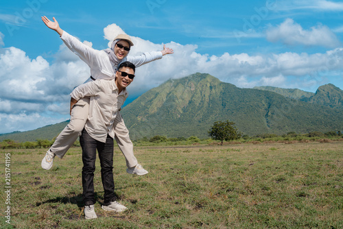 A joyful couple share a playful piggyback in a sunlit grassy plain with a dramatic Baluran mountain backdrop, wearing sunglasses and casual clothes while enjoying travel, freedom and outdoor adventure