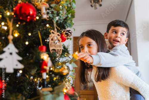 Obraz na plátně Kids decorating christmas tree, brother on sister's back, enjoying festive holid