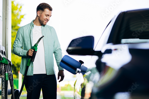 Man holding a green fuel nozzle, filling his car with gasoline at a modern service station, looking down with a smile