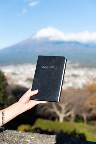 Bild auf Leinwand A hand holds a black, hardcover Holy Bible aloft with the iconic snow-capped Mount Fuji and Fujinomiya city in the background, from the Shirao Mountain Park (Shiraosan), Japan