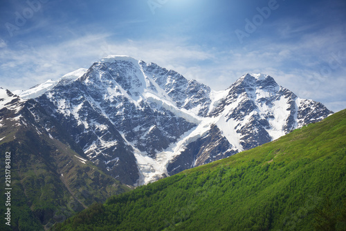 Landscape of a high rocky mountain with massive snowy peaks and glacier slopes under a blue sky with soft white clouds.