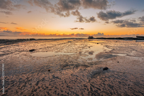 A large wave cut rock platform at low tide serves as a foreground across the sea to the orange glow of sunrise behind clouds on the horison on the coast at Burrill Lake in New South Wales, Australia.