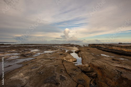 A wave cut platform of rock, the various cracks and inlets in the rock, and even the clouds, lead the eye to an enormous thunder cloud in the distance at Burrill Lake, New South Wales, Australia.