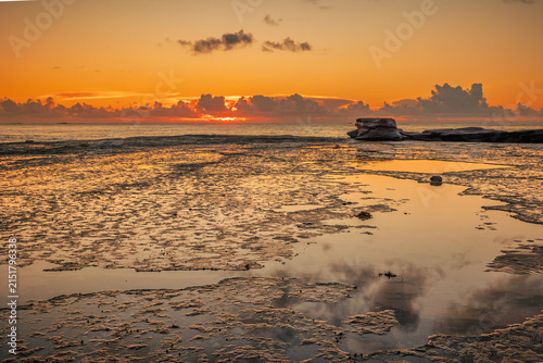 Clouds on the horison look like they are on fire at sunrise as viewed across a flat, wave cut platform with reflections in the puddles on the coast at Burrill Lake in New South Wales, Australia.