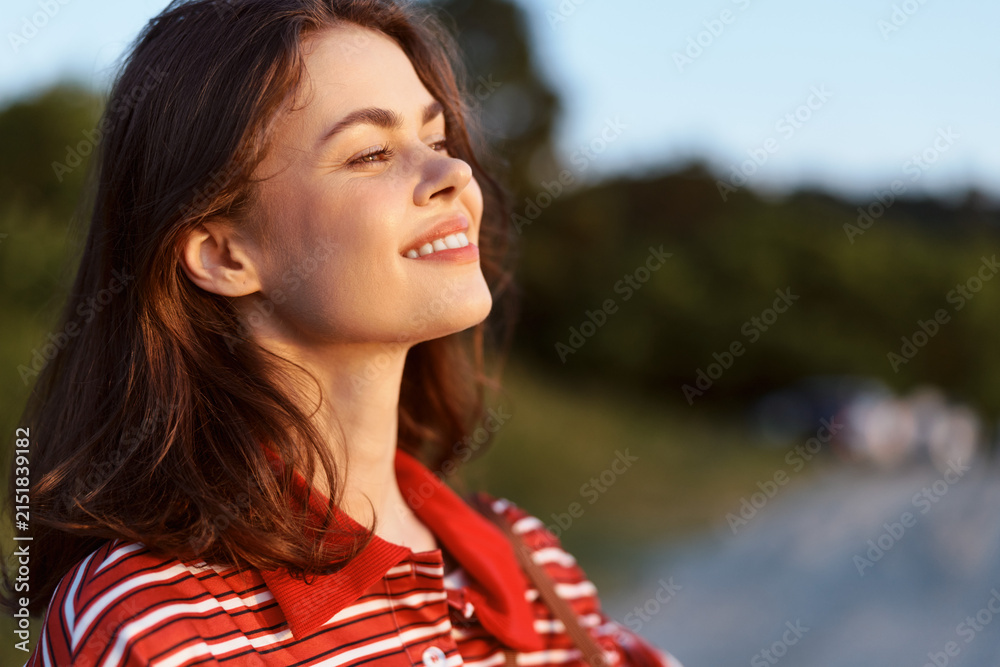 Obraz premium Young woman in a red striped shirt smiles softly, eyes closed, basking in sunlight outdoors. A natural light portrait perfect for lifestyle, travel, or wellness concepts.