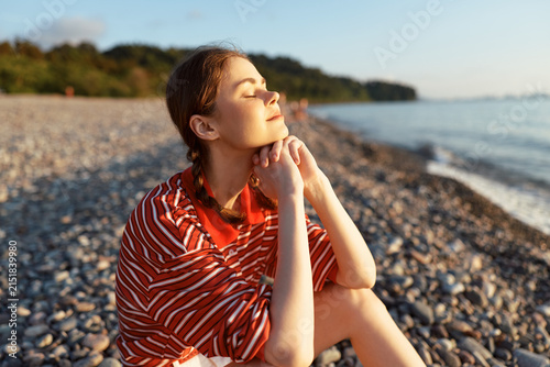 Photography A woman sits on a pebbled beach in a red striped shirt, eyes closed, basking in the sun