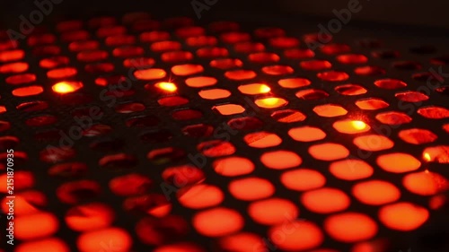 Glowing orange light emitting from behind a dark metal mesh grid. Abstract industrial texture with perforated holes and warm illumination for technology concept.