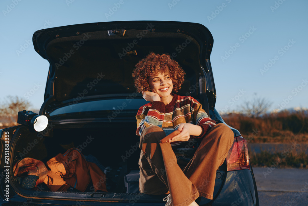 Obraz premium Girl child car trunk smile sunset outdoors lifestyle, sitting on open trunk at dusk, relaxed casual pose, warm golden light, with curly hair playful expression family road trip stop outdoors.