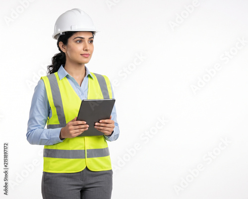 young Indian female professional wearing a white safety helmet and high visibility vest holding a black tablet in a confident business success pose with clean copy space