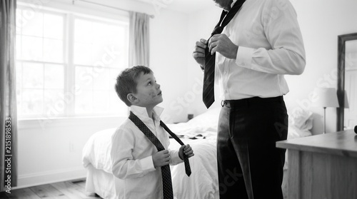 Young boy looks up at his father while they both put on neckties in a bright bedroom during morning routine. This black and white portrait captures a tender and heartwarming moment of