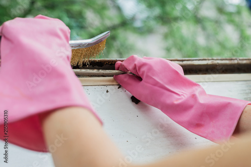 Close view of hands in pink rubber gloves using wire brush to clean rusty metal window frame before painting, with peeling paint, corrosion flakes, worn surface, and blurred green outdoor background
