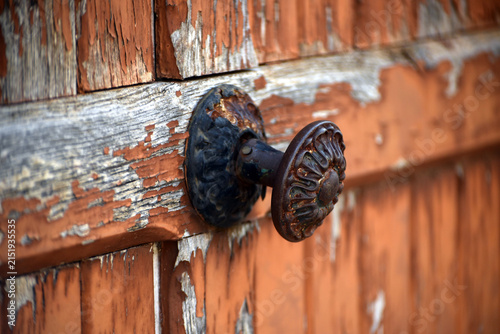 Close up of an old rusty door handle attached to a weathered brown wooden door with peeling paint, showing a rustic architectural detail full of character.

