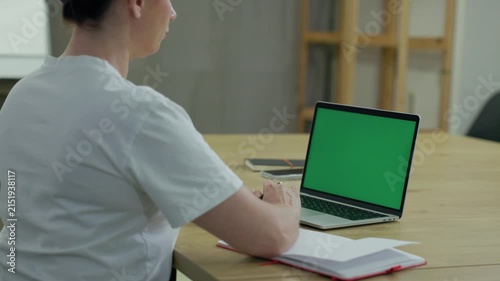 Woman Working On Laptop Computer with Green Mock Up Screen At Home Office Space. Female Freelancer Browsing Green Laptop Screen Writing Information in Notebook