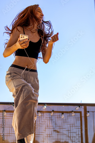 Red haired Gen Z teenage girl jumps joyfully with arms raised high while dancing on sunny balcony. Concept of pure happiness and energetic self expression through dance in open air.