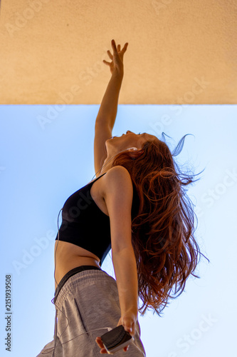 Red haired Gen Z teenage girl dances with arm raised and hair flying against clear blue sky. Concept of dynamic self expression and carefree movement capturing youthful energy outdoors.