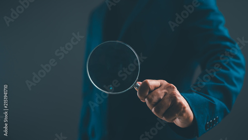 Fotografie A person in a blue suit holds a magnifying glass, focusing on the viewer against a dark background