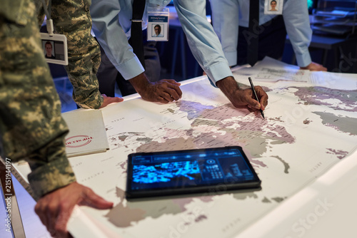 Diverse military officers and government officials analyzing world map on table during strategic planning session, suitable for national security and defense industry marketing
