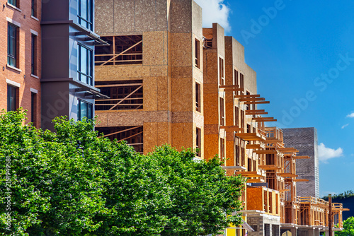 Modern multi-story wood frame building under construction with OSB panels in an urban setting, featuring green trees and a clear blue sky.