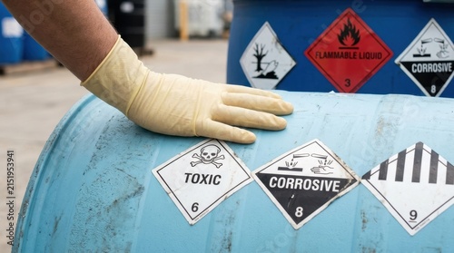 Professional worker hand wearing protective yellow glove touching industrial blue chemical barrel with toxic and corrosive hazard warning signs labels in a warehouse setting storage