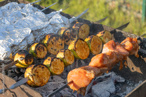 Delicious shish kebab and zucchini grilling on skewers over hot coals in a backyard grill. Meat and vegetables cooking outdoors during a sunny weekend picnic, preparing a tasty meal for family