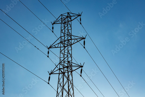 High voltage electrical transmission tower and power lines silhouetted against a clear blue sky during a bright, sunny day, with strong backlight.