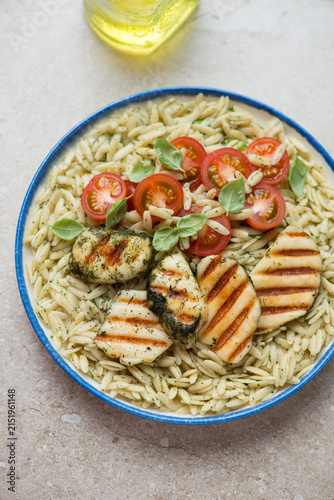 Plate of orzo with basil oil, cherry tomatoes and grilled halloumi on a beige stone background, vertical shot, elevated view