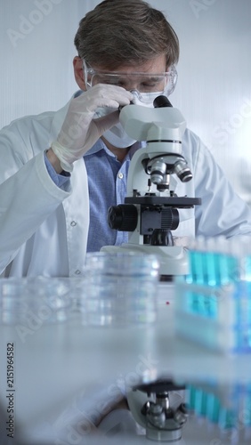 Male scientist researcher wearing lab coat, white protective gloves, medical mask and glasses, is using microscope in modern laboratory setup, portrait view. Low key lighting photo. Science