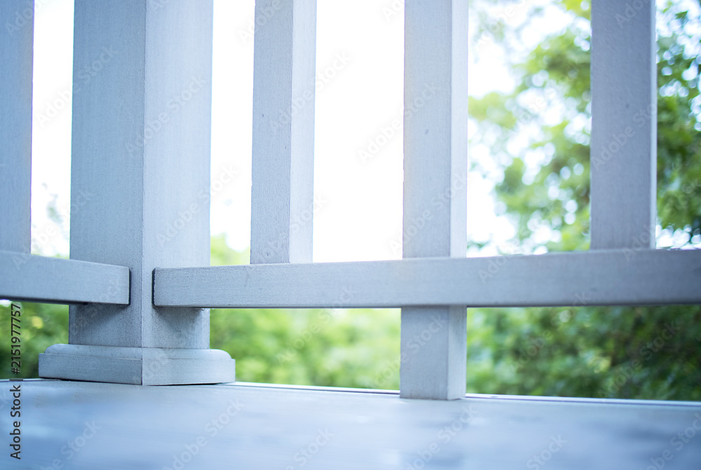Fototapeta premium Close-up view of wooden balcony railing with vertical slats and horizontal support, overlooking green foliage and trees in a bright outdoor setting