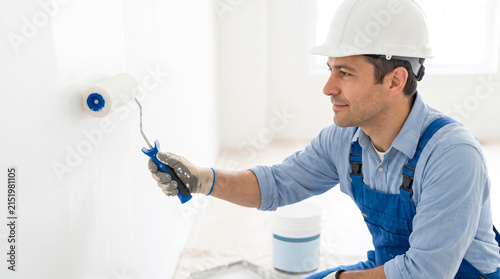 A man is painting a wall with a roller inside a room. He is wearing a hard hat and working diligently. The room is empty and has bright light coming through windows