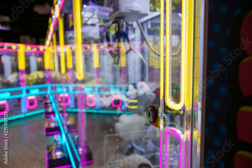 Close-up captures the vivid neon framing in yellow, pink, and cyan that outlines the claw machine's glass and structure; blurred background reveals stacked plush prizes and mechanical components, emph