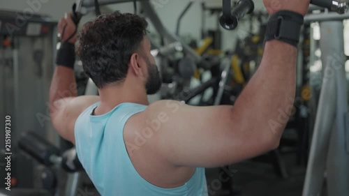 Indian Asian fit young man or male using a lat pulldown machine, performing resistance weight training exercise or workout targeting the back and shoulders in modern gym. Fitness, bodybuilding concept