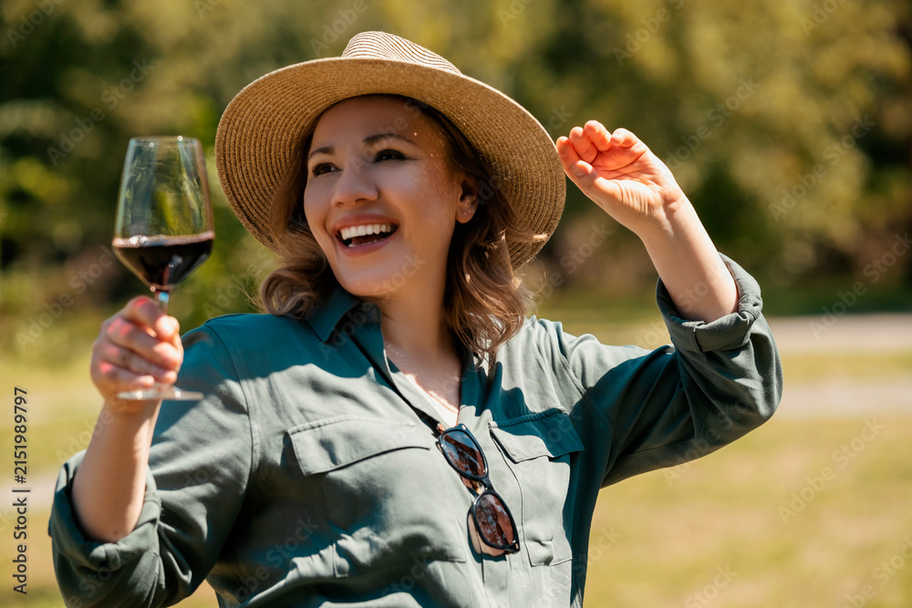 Obraz premium Happy smiling woman in straw hat holding glass of red wine outdoors in sunny park. Cheerful mood and joyful summer picnic vibe.