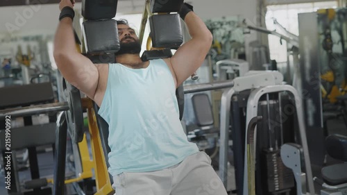 A young Indian Asian fit muscular male performs a leg squat exercise using a machine as part of resistance or weight training in a modern gym. Fitness, workout, healthy lifestyle, wellness concept