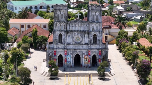 Cinematic drone footage using a 3x telephoto zoom facing the main facade of Mang Lang Church in Phu Yen, Vietnam. The camera gradually pulls backward while tilting upward,