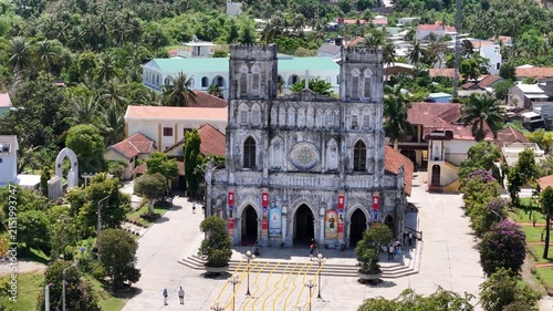 Cinematic drone footage using a 3x telephoto zoom, orbiting around Mang Lang Church while gradually descending toward its main facade in Vietnam. The smooth movement brings the historic Catholic Churc
