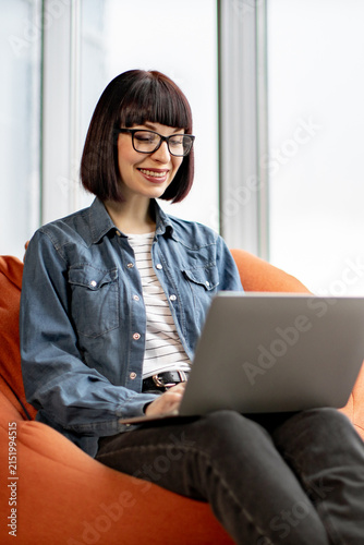 A young woman with glasses and a bob haircut smiles while working on her laptop computer, seated comfortably on an orange beanbag chair